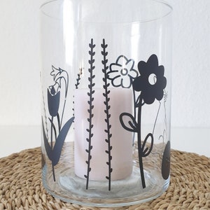 Puede incluir: Un portavelas de vidrio transparente con un dise&ntilde;o floral negro. El dise&ntilde;o presenta un tulip&aacute;n, una margarita, una flor grande y hierba. Una vela blanca est&aacute; dentro del portavelas.