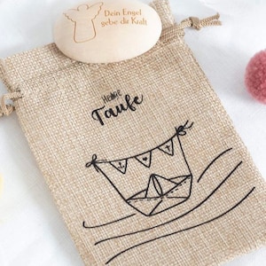 May include: A small burlap bag with a wooden angel charm inside. The bag has a black and white illustration of a boat with a banner that says "Heilige Taufe". The angel charm has the text "Dein Engel gebe dir Kraft" engraved on it.