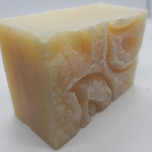 May include: A light yellow, rectangular bar of handmade soap. The soap has a textured surface and a carved design on one side. The cut edge reveals a slightly uneven texture. The soap appears to be freshly made.