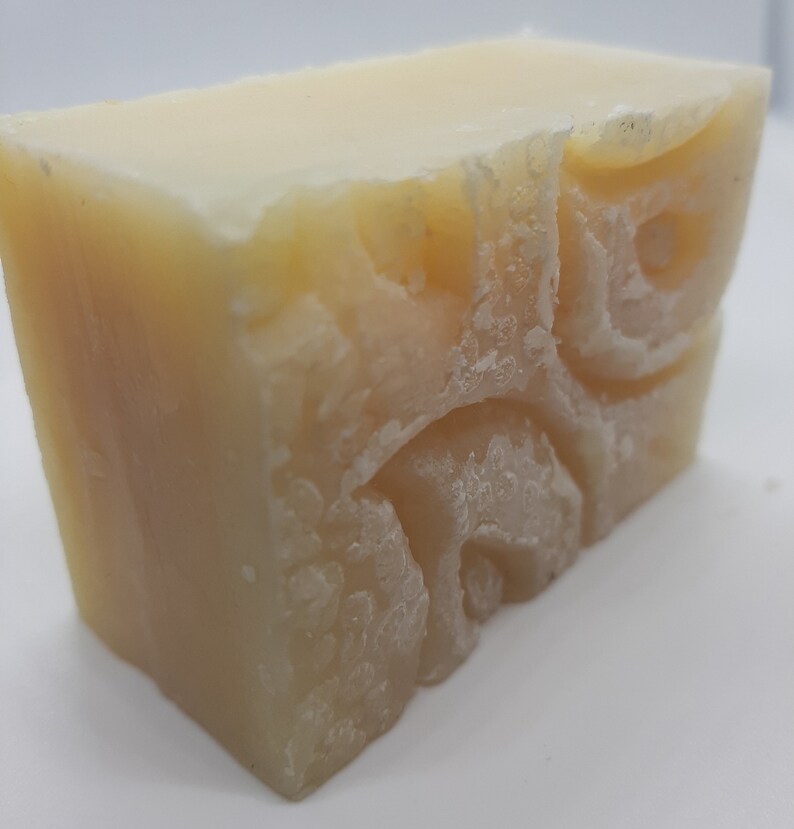 May include: A light yellow, rectangular bar of handmade soap. The soap has a textured surface and a carved design on one side. The cut edge reveals a slightly uneven texture. The soap appears to be freshly made.