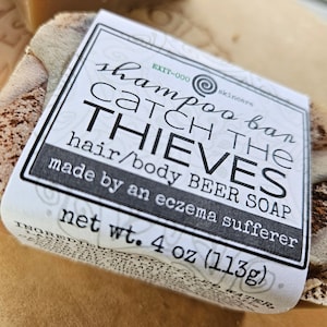 May include: A white label with black text reads "shampoo bar CATCH THE THIEVES hair/body BEER SOAP made by an eczema sufferer net wt. 4 oz (113g)". The label is attached to a brown and green bar of soap.