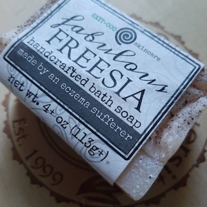 May include: A white bar of soap with a label that reads "fabulous FREESIA handcrafted bath soap made by an eczema sufferer net wt. 4+ oz (113g+) EXT-000 skincare Est. 1999".