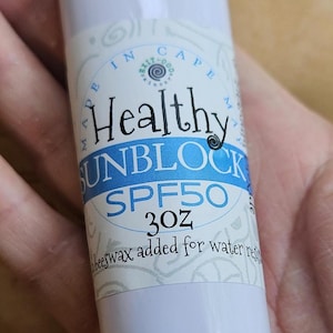 May include: A white tube of Healthy UNBLOCK SPF50 sunscreen. The label includes the text "Healthy UNBLOCK SPF50 3oz beeswax added for water res". The tube is held in a hand.