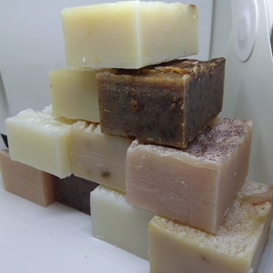 May include: A stack of handmade soap bars in various colors and textures. The soaps range from light beige to dark brown, with visible natural ingredients. The bars are rectangular and arranged in a visually appealing pyramid shape, showcasing their artisanal quality.