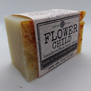 May include: A bar of handcrafted bath soap labeled "FLOWER CHILD" made by an eczema sufferer. The soap is a light beige color with a yellow tint and is packaged in a white label with black text. The label also includes the text "EXIT-000 skincare" and "net wt. 4 oz (115g)".