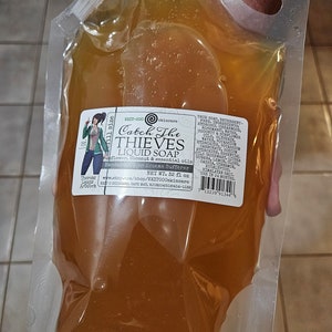 May include: A transparent pouch containing amber liquid soap. The label reads "Catch The Thieves Liquid Soap" and includes a cartoon illustration. The pouch has a white spout for dispensing. The label also lists ingredients.