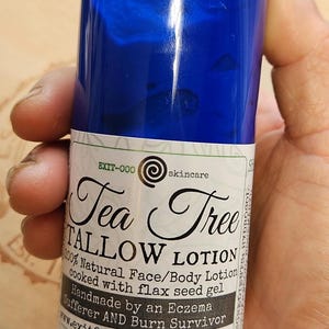 May include: A blue bottle of Tea Tree Tallow Lotion, a 100% natural face and body lotion made with flax seed gel. The bottle is labeled "Tea Tree Tallow Lotion" and "Handmade by an Eczema Sufferer AND Burn Survivor". The bottle is 8 fl. oz.