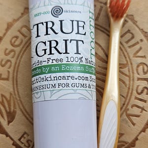 May include: A white tube of toothpaste with the text "TRUE GRIT TOOTH" and "Fluoride-Free 100% Natural Made by an Eczema Sufferer" on the label. The tube is next to a gold toothbrush with red bristles.