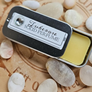 May include: A small, open, rectangular metal tin of solid perfume labeled "Tuberose Solid Perfume." The perfume is a light yellow color. The tin is surrounded by smooth, white and tan stones on a brown surface.