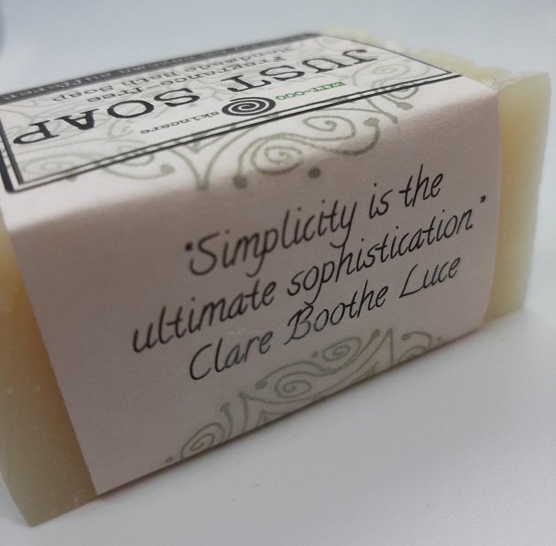 May include: A rectangular bar of handmade soap with a pale colour. The soap is wrapped with a light pink label. The label features the text "Simplicity is the ultimate sophistication" by Clare Boothe Luce, along with the words "JUST SOAP" and "fragrance-free".