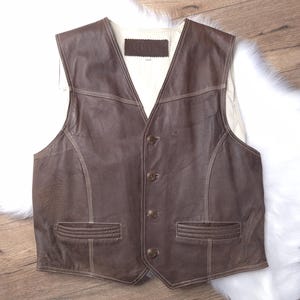May include: A brown leather vest with a button-up front and two pockets. The vest has a light brown stitching detail.