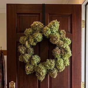 May include: A circular wreath composed of numerous dried green hydrangea blossoms, mounted on a dark brown wooden door with a brass handle and lock mechanism. The wreath features a dense, textured arrangement of spherical flower clusters.