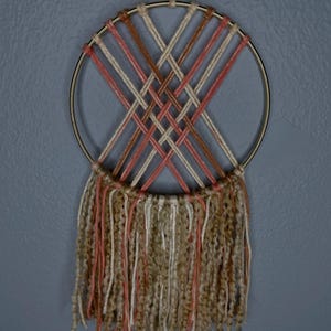 May include: A woven wall hanging featuring a gold metal ring with a woven pattern in shades of pink, brown, and cream. The bottom of the ring has long, textured fringe in matching colors. A loop of string is attached for hanging.