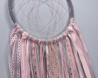 May include: A gray and pink dream catcher with a circular hoop and a variety of pink and gray ribbons hanging from the bottom.