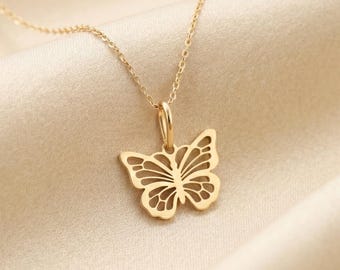 May include: A gold-colored butterfly pendant necklace. The butterfly pendant has a detailed, openwork design. The necklace chain is delicate and thin. The background is a soft, neutral fabric.