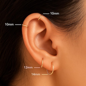 May include: Four thin gold hoop earrings worn in the ear. Two hoops are 10mm in diameter, one hoop is 12mm in diameter, and one hoop is 14mm in diameter. The earrings are simple, circular bands of gold metal positioned in the earlobe and cartilage.