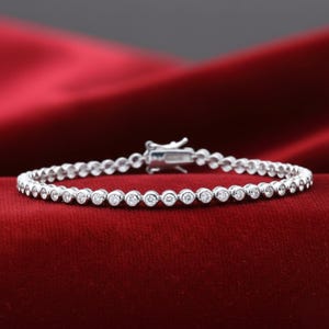 Puede incluir: Una delicada pulsera de plata con numerosas piedras preciosas pequeñas, redondas y transparentes. La pulsera se muestra sobre un fondo rojo oscuro texturizado y tiene un cierre seguro.