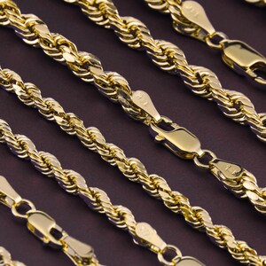 May include: Five gold rope chains of varying lengths and thicknesses. The chains are laid out on a dark purple surface.