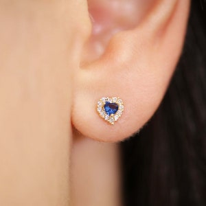 K&ouml;nnte beinhalten: Ein goldfarbener herzf&ouml;rmiger Ohrstecker mit einem blauen Mittelstein, umgeben von einem Kranz aus kleinen, klaren Steinen. Der Ohrring befindet sich im Ohr einer Person.