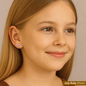 K&ouml;nnte beinhalten: Ein junger Mensch mit blonden Haaren tr&auml;gt herzf&ouml;rmige Ohrstecker. Die Ohrringe haben einen rosa Mittelstein, der von kleinen, klaren Steinen umgeben ist. Der Text "July Ruby Shown" ist unten rechts sichtbar.