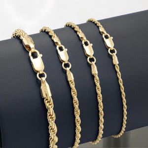May include: Four gold-colored rope chains with lobster claw clasps are displayed against a dark blue background. The chains vary slightly in thickness, showcasing a classic jewelry design. The chains are made of a shiny, reflective material.