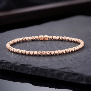 Puede incluir: Una pulsera de cuentas en tono oro rosa con pequeñas cuentas esféricas texturizadas dispuestas en una banda circular continua. Dos eslabones rectangulares centrales están estampados con el texto 925 e ITALY, indicando composición y origen.