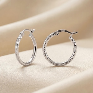 May include: A pair of silver hoop earrings with a textured, diamond-like pattern. The earrings are displayed on a beige, satin-like surface. The clasp of one earring is marked with "925".