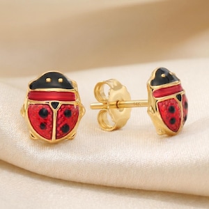 May include: A pair of stud earrings shaped like ladybugs with red and black enamel detailing on a gold-tone base. The earrings feature a post back with a butterfly clutch, and the post is clearly stamped with the text 14K for gold purity.