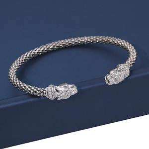 May include: Silver bracelet with a textured, woven design. The bracelet has two panther head terminals, each decorated with small, sparkling gemstones. The bracelet is set against a dark blue background.