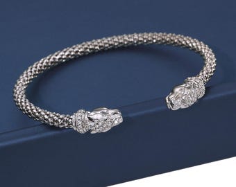 May include: Silver-toned bracelet with a textured, woven design. The bracelet features two panther head terminals, each adorned with small, sparkling gemstones. The bracelet is set against a dark blue background.