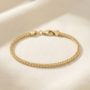 May include: A gold-colored bracelet with a woven chain design. The bracelet features a lobster clasp closure and is displayed on a cream-colored fabric. The bracelet is approximately 7 inches in length.