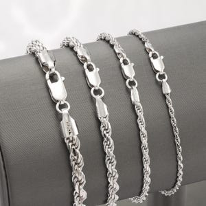 May include: Four silver-colored rope chain necklaces with lobster claw clasps are displayed against a gray background. The chains vary in thickness, showcasing different styles of jewelry. The clasps are shiny and reflect the light.