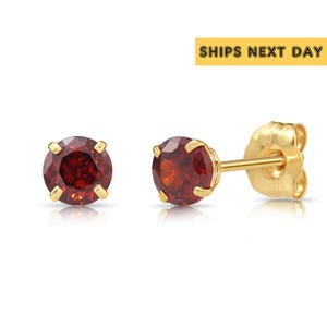 May include: A pair of gold stud earrings with round, deep red gemstones. The earrings have a classic design with a four-prong setting. The image includes the text "SHIPS NEXT DAY".