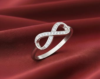 Puede incluir: Un anillo de tono plateado con un símbolo de infinito en el centro adornado con pequeñas piedras claras y brillantes. La banda es lisa y pulida, con el sello 925 claramente grabado en la superficie interior del aro metálico del anillo.