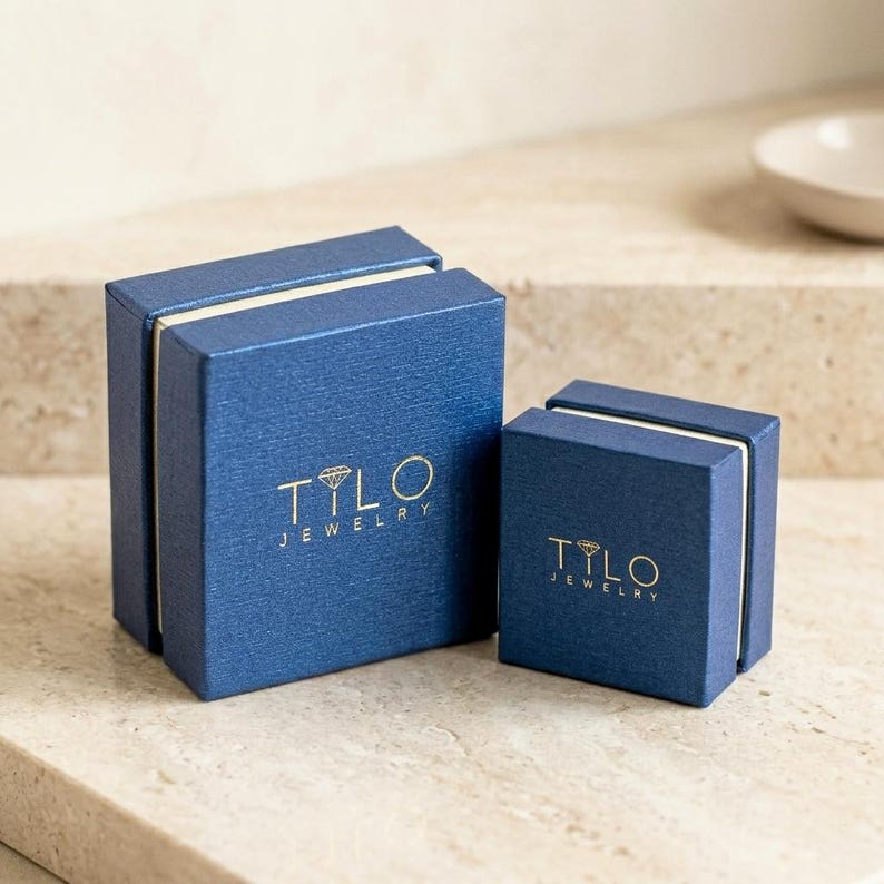 May include: Two square blue jewelry boxes with a textured finish. Each box features the gold-colored text TILO JEWELRY and a small diamond icon printed on the lid. The larger box is positioned to the left of the smaller, matching square box.
