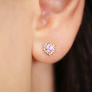 K&ouml;nnte beinhalten: Nahaufnahme eines herzf&ouml;rmigen Ohrsteckers in Gold. Der Ohrring hat einen hellvioletten herzf&ouml;rmigen Mittelstein, der von einem Kranz aus kleinen, klaren Kristallen umgeben ist. Der Ohrring ist an der Haut des Ohrs einer Person befestigt.