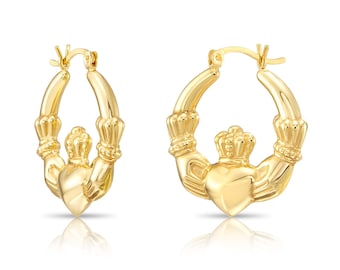 Puede incluir: Un par de pendientes de aro Claddagh dorados. Cada pendiente presenta un diseño de corazón, corona y manos. Los pendientes se muestran sobre un fondo blanco, con un ligero reflejo debajo.