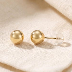 May include: A pair of gold ball stud earrings with a polished, reflective finish. Each earring features a smooth, spherical gold bead attached to a thin post, secured with a clear, translucent plastic earring back for comfortable wear.