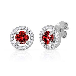 May include: A pair of silver stud earrings with a red gemstone surrounded by a halo of smaller white gemstones.