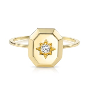 May include: A gold ring featuring an octagonal face with a starburst design and a central, round, clear gemstone. The ring's band is thin and smooth, and the ring is set against a white background.