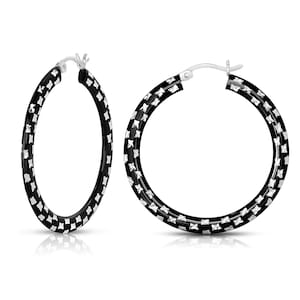 May include: Two hoop earrings. The earrings are black with a silver-colored pattern. The earrings are round and have a clasp closure. The earrings are displayed on a white background.