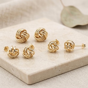 May include: Six gold-colored knot stud earrings displayed on a light-colored, square surface. The earrings are arranged in a group, showcasing their intricate, intertwined design. The earrings have a polished, shiny finish, reflecting the light.