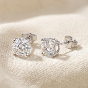 May include: A pair of silver-toned stud earrings featuring round, brilliant-cut clear stones held in four-prong settings. Each earring includes a matching metal post and a secure butterfly-style backing, showcasing a classic and reflective design.