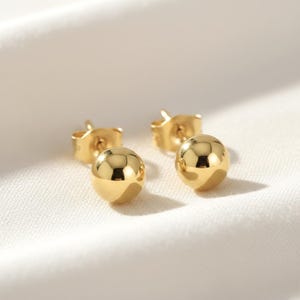 May include: A pair of polished gold-tone ball stud earrings. Each earring features a smooth, spherical bead design with a reflective metallic finish and a standard post backing, showcasing a simple and clean aesthetic for everyday wear.