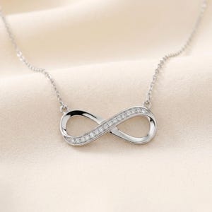 May include: A silver necklace with an infinity symbol pendant. The pendant is set with small, clear gemstones. The chain is fine, and the pendant is displayed against a soft, cream-coloured backdrop. The necklace is a piece of jewellery.