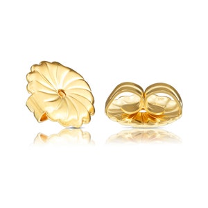 May include: A pair of gold-toned earring backs. The backs are designed to be worn on the back of an earring to secure it to the earlobe. The backs have a circular shape with a small hole in the centre.