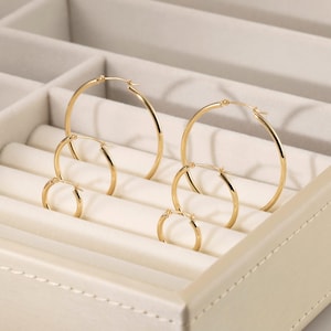 Può includere: Una collezione di orecchini a cerchio dorati di varie dimensioni, esposti in un organizer per gioielli color crema. Gli orecchini sono disposti in coppie, mostrando il loro design circolare semplice e la finitura lucida. L'organizer ha più scomparti.