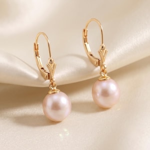 May include: A pair of gold leverback earrings, each featuring a single round, pale pink pearl suspended from a polished gold setting. The earrings have a secure hinged closure mechanism and a smooth, lustrous finish on both the metal and pearls.