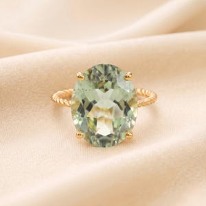 May include: A gold ring featuring a large, oval-cut, pale green gemstone. The gemstone is set with four prongs and the band has a twisted rope design. The ring is displayed on a cream-colored fabric.