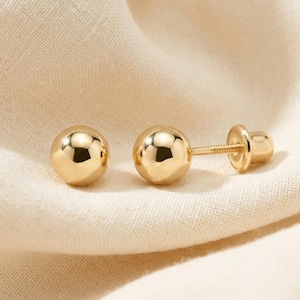 May include: A pair of gold ball stud earrings. The earrings have a polished, reflective surface and a screw-back closure. The earrings are displayed on a cream-colored fabric.